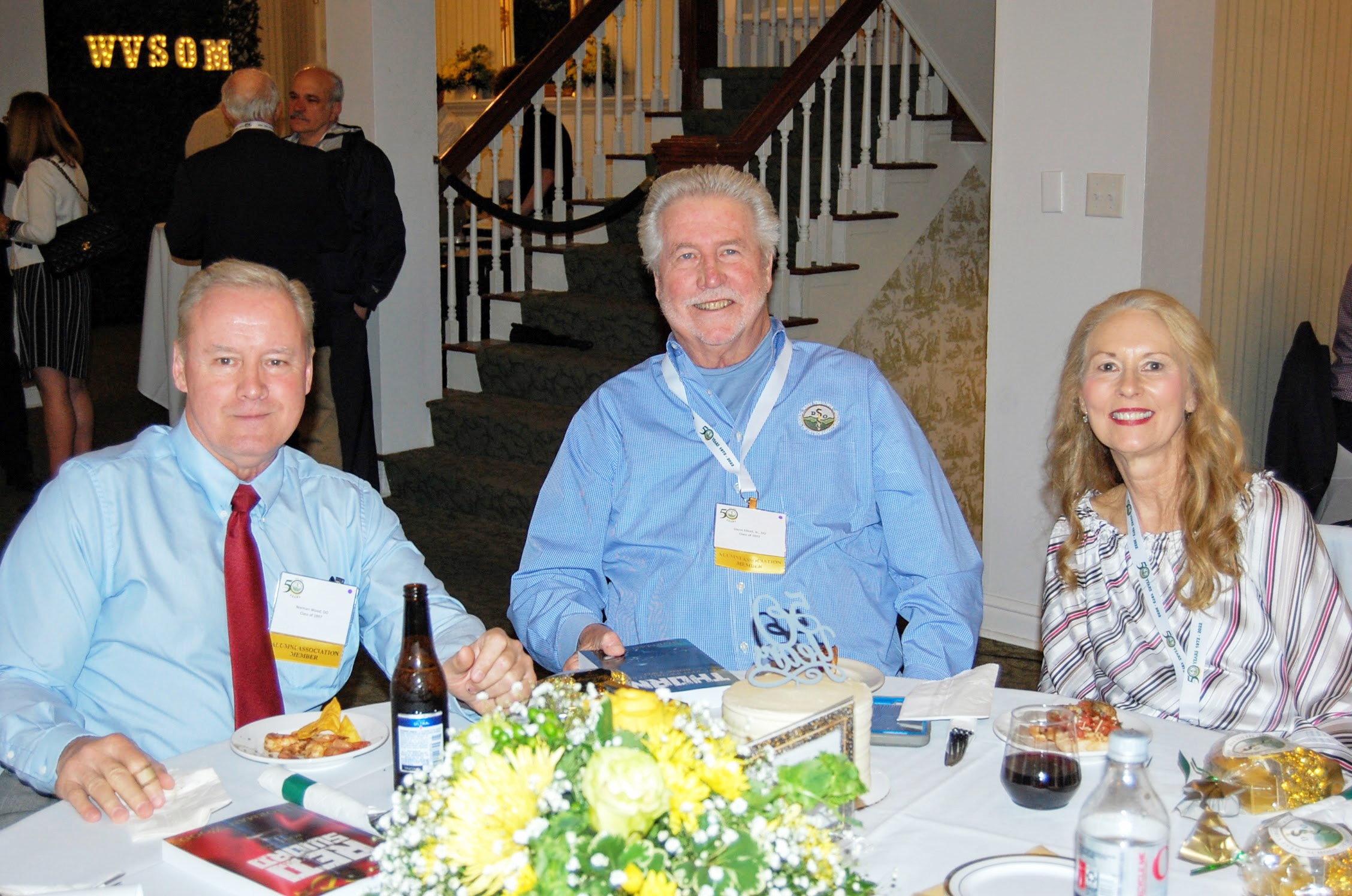 Three attendees seated at table with food and drinks smile for camera, WVSOM in lights sign can be seen in the background