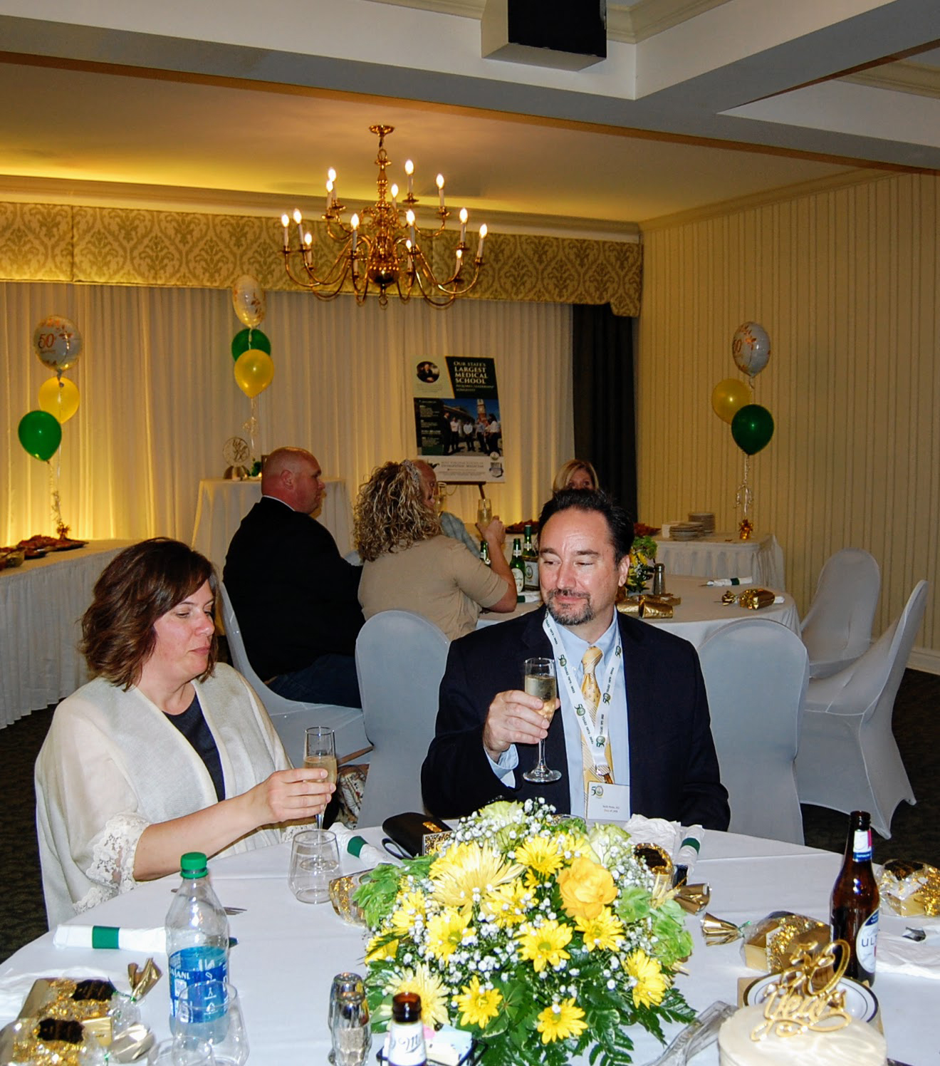 Man and woman appear to be starting a champagne toast, another table can be seen in the background also holding up glasses
