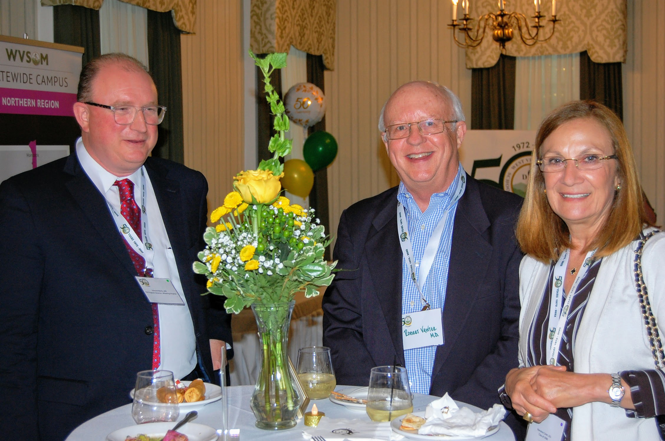 Two men, one name tag reads Robert Vanter, M.D., and woman smile for camera while standing at refreshement table with flowers 