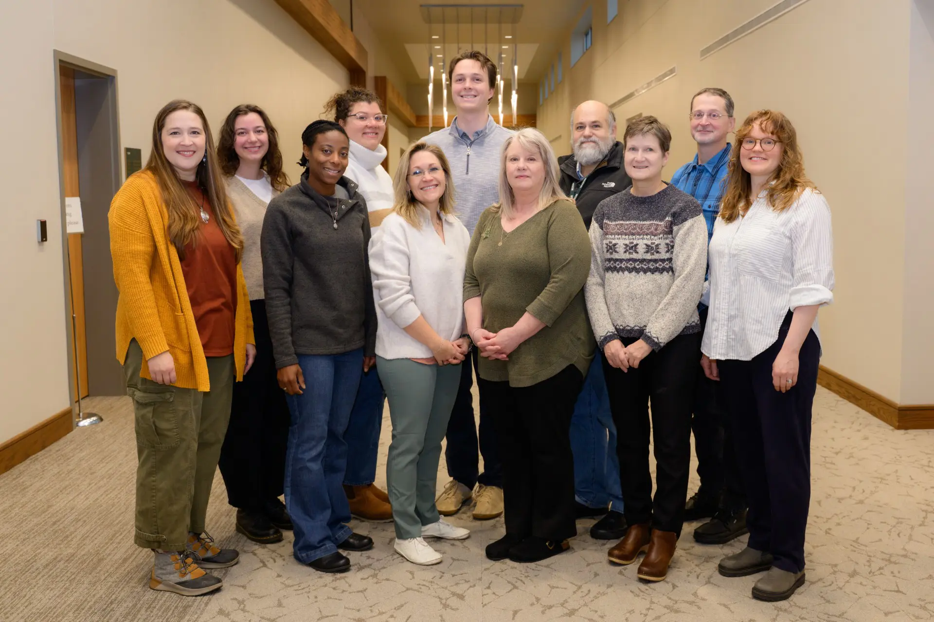 Eleven people stand in two rows in tall ceilinged hallway.