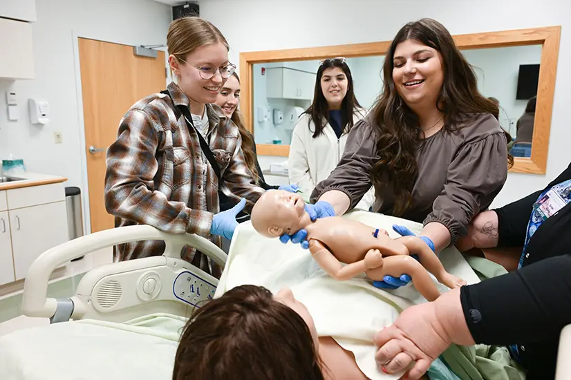 Students around a humanoid simulation robot, one holds out simulation baby robot to simulation mother.