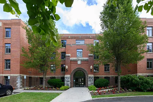 Brick building showing architectural block base, protruded arched entry with multi direction walkway leading to and around the building. Two Large trees on either side of the entrance, with other smaller landscaping.
