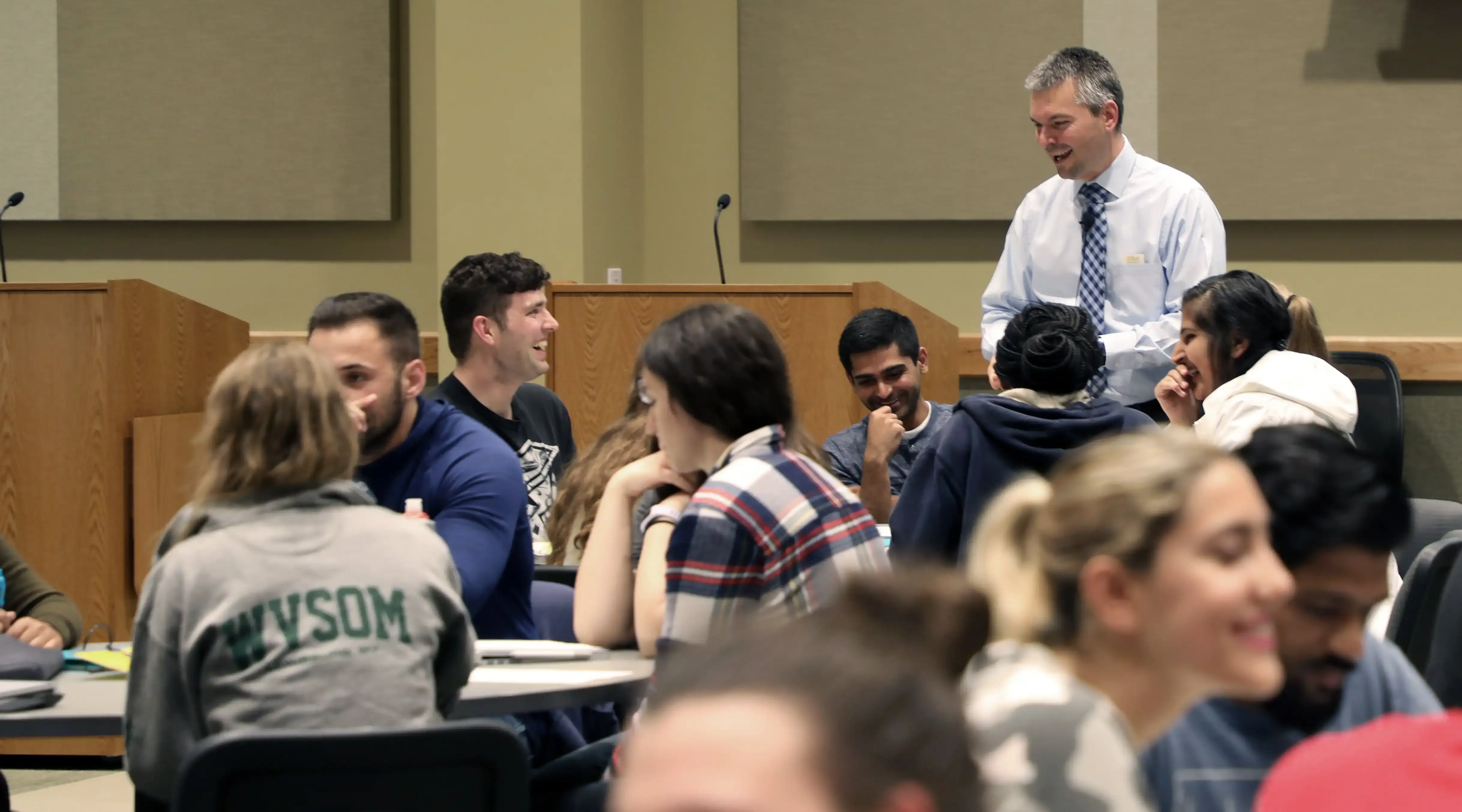 Professor speaking to classroom of students sitting at round tables, student seen smiling and laughing