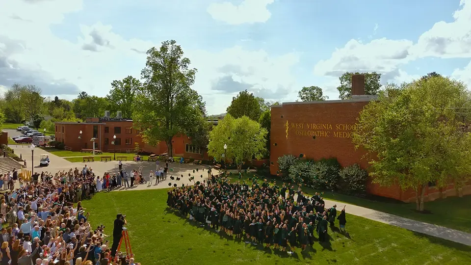 Graduates in cap and gowns standing in a group outside crowd gathered around them. Photographer takes picture of graduates from a ladder.