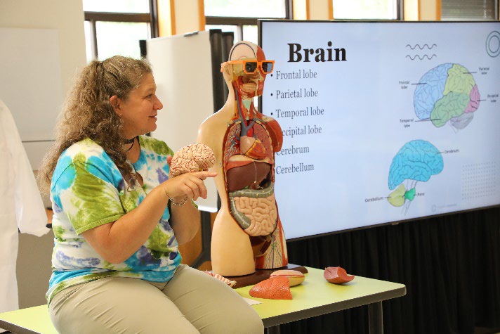 Karen Wines instructucts students from desk with anatomy torso and head showing exposed organs and wearing sunglesses